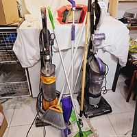 Full view of vacuum cleaners and cleaning tools including Dyson DC29, Dirt Devil, Swiffer floor and wet mop, giant mop, broom, and cleaning pads.