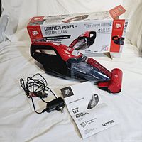 Dirt Devil cordless handheld vacuum with charger and user manual alongside original product box shown on white fabric background.