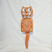 Full view of orange Kit-Cat Klock with rhinestones on eyes, bow tie, ears, paws, and tail. Clock face shows time and cat tail pendulum attached.