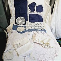 Overview photo showing assorted crochet doilies, navy blue linen pieces with crochet trim, white embroidered napkins, and other handmade textiles laid out on a white sheet.