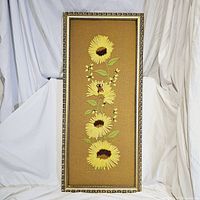 Full view of framed vintage crewel stitchery tapestry featuring sunflowers, daisies, and ladybug on tan fabric background, standing against white backdrop.