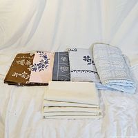 Six vintage tablecloths folded showing various colors and embroidery patterns including brown, pink with blue, gray with white, white with blue, and a woven floral pattern.