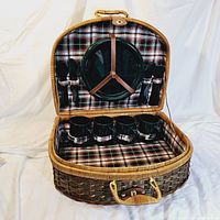 Open wicker picnic basket showing four dark ceramic plates on lid secured with leather straps, and four dark mugs with plaid holders fitted in base compartment. Utensils with black handles held in elastic loops on lid.