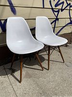 Two white Eames style chairs with wooden Eiffel tower legs positioned outdoors showing front and side views.