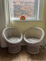Pair of white rattan barrel chairs positioned in front of a window with wood flooring.