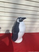 Photo of front-left side of the blow mold penguin showing painted black and white body, standing on red cloth against siding background.
