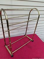 Side angled view of brass tone bamboo style towel rack showing arched vertical supports and horizontal drying bars on red surface with siding background.