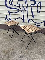 Two Ikea Tarno folding patio cafe tables with wooden slatted tops and black folding metal legs standing outside against a wall with graffiti.