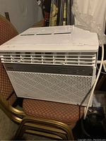 Front view of white Haier window mount air conditioner on stacked chairs, showing vented front grille and control panel.