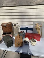 Overview of all items on a white table including metal and wooden boxes, ceramic pots, a large clear oiler bottle, metal oil can, and decorative plaque