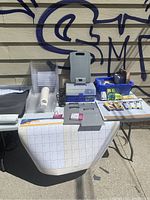 Overall lot showing clipboards, planner calendar, plastic trays, lidded storage box, pens, markers on a folding table outdoors.