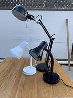 Three office/task lamps shown together on a wooden table outdoors, including two black adjustable lamps and one white lamp with flexible neck.