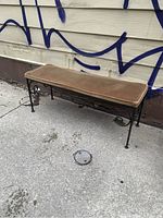 Full view of a black wrought iron bench with brown velvet cushion on a concrete surface against a wall with blue graffiti.