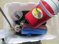 Photo showing multiple camping items inside clear plastic bin, including fishing pole, blue and yellow camping mat, small propane heater, silver camping stove or lantern component, and red Coleman water jug.