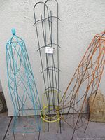 Photo showing three garden trellises: two cone-shaped wireframe trellises in blue and orange; one black and yellow metal stake trellis with circular rings for plant support.