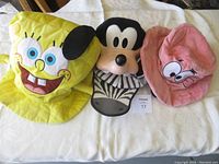 Four character hats laid out, from left to right SpongeBob SquarePants in bright yellow, Goofy hat with plastic face and black and white zebra pattern fabric, pink Patrick Star hat with embroidered eyes.