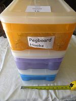 Stack of three plastic organizer drawers with orange, purple, and blue color accents. Label on top drawer reads 'Pegboard Hooks'.