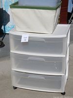 Three-drawer white plastic storage organizer with translucent drawers and wheels. Beige fabric bin rests on top.