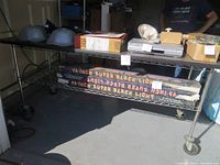 Full side view of large metal rolling work cart table showing long flat top, wire lower shelf, and wheels. Boxes and items on top are not included in sale.