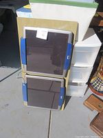 Two dark rectangular solar panels mounted on wooden boards with blue handles on the sides, and a white carrying case behind them.