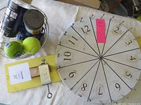 Photo of metal spinning wheel game numbered 1 to 12 with cotter pin stopper alongside a basket containing assorted balls and plastic containers.