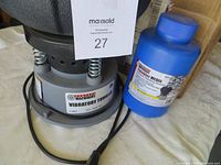 Gray vibratory tumbler with power cord and blue container of tumbler media in photo, showing brand label.