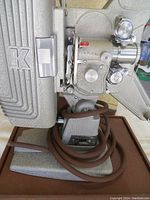 Side view of the film projector showing brand 'K', knobs, lens, and power cord coiled on base