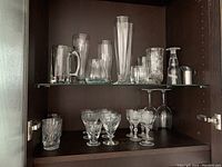 Two shelves inside a cabinet holding various crystal and glass barware including beer glasses, tumblers, wine glasses, and a small bottle with stopper.