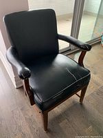 Black faux leather armchair placed on wooden floor near glass door, showing front and top views with visible cracks on seat upholstery and worn armrests.