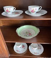Two porcelain cups and luncheon plates with pink and yellow floral pattern on white porcelain with ridged edges on shelf above