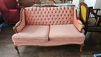 Front view of pink tufted upholstered wooden framed loveseat showing full furniture item with cushions and carved legs.