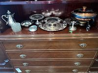 Wide shot showing multiple silver plated items on a wooden dresser, including teapot, trays, candy dishes, and copper chafing dish with glass lid on burner stand.