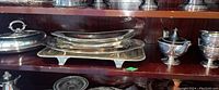 Shelf showing group of silver plated serving trays, covered dish, coffee pot, sugar bowl, and creamer.