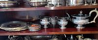 View of silver plate trays, teapot, sugar bowl, creamer, and wine bottle stand arranged on shelf