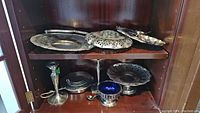Silverplate candlestick holder, lidded sugar bowl, round pedestal cake tray, salt cellar with spoon, flower frog with blue interior, round tray with pierced edge, and smaller oval tray with decorated edge displayed on wooden shelves.