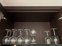 Shelf with 12 wine glasses: 10 matching with identical shape and size, 2 different wine glasses. All clear glass with stems and bowls typical of wine glasses.