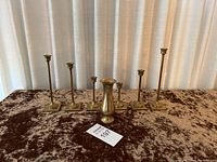 Photo showing five metal taper candle holders of varying heights and one small metal vase on a brown velvet surface with cream curtains background.