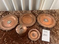 Full view of three copper plates, small hammered copper container with brass knob, and small copper ashtray arranged on brown fabric surface.
