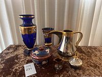 Photo showing six decorative items on a table: two blue and gold vases, a silver and gold coffee pot, a covered floral lidded container, a small floral creamer, and a silver and gold urn-shaped container.