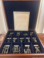 Full view of the open wooden display case with 15 pewter figurines arranged in individual slots lined with blue velvet, with a labeled chart inside the lid.