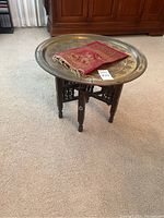 View of folding side table with ornate brass tray top and small red textile on top, showing side and top profile of the table.