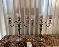 Two silver plated candelabra displayed on brown velvet surface with white curtains background. Each has three arms and a central candle holder with textured glass candle holders containing red candles.