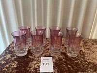 Set of eight cranberry flash thumbprint iced tea glasses arranged in two rows on a brown velvet surface in front of a cream curtain.