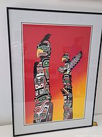 Framed artwork showing two totem poles on a red to yellow gradient background, with black frame and white mats.