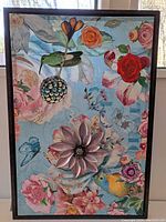 Full frontal image of the large mixed media collage framed artwork with colorful flowers, birds and butterfly on a blue background and a 3D metal daisy flower attached.