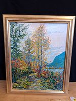 Full view of the framed oil painting showing landscape scene with river bank, trees, and mountains in autumn colors.