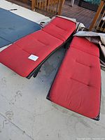 Two red lounge chairs with black resin wicker frames and matching red cushions laid out on a concrete surface. Cushions have button tufting and covers included.