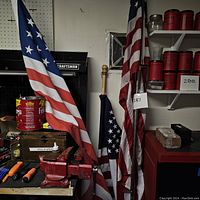 Three American flags attached to poles standing vertically, showing stars and stripes; background includes workshop items and shelves with red containers.