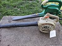 Two handheld garden blowers, green electric Weed Eater and beige gas-powered models, both with air tube attachments, placed on concrete outdoors.