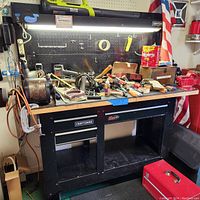 Full front view of the Craftsman black metal storage workbench with assorted tools on top, pegboard with hooks mounted on the back, illuminated light bar overhead, red metal toolbox on floor.
